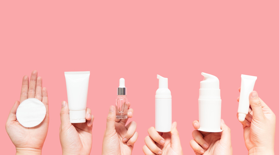Três pares de mãos segurando diferentes embalagens de cosméticos em um fundo rosa.