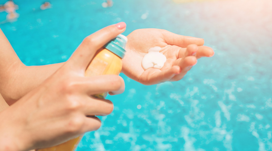 Plano detalhe duas mãos em frente a uma piscina, uma delas segurando o tubo de protetor solar e a outra com produto depositado.